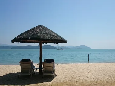 Tropical beach scene with two lounge chairs under a thatched umbrella, overlooking calm blue sea and distant islands.
