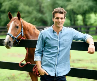 A man in a denim shirt leans on a fence beside a horse in a lush green field.