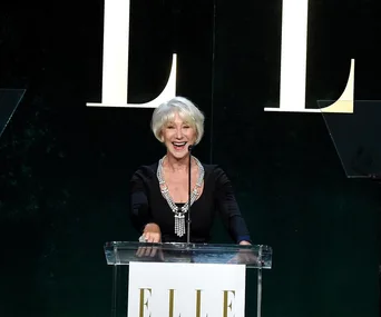 Helen Mirren speaking at a podium on stage during an Elle event, with a large "E" in the background.