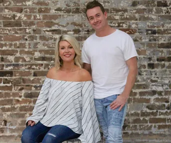A couple smiling at the camera, the woman is seated in a striped top and jeans, the man stands in a white shirt and jeans.