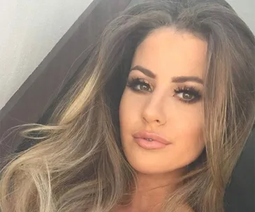 A woman with long, highlighted hair and makeup is taking a selfie, looking into the camera with a neutral expression.