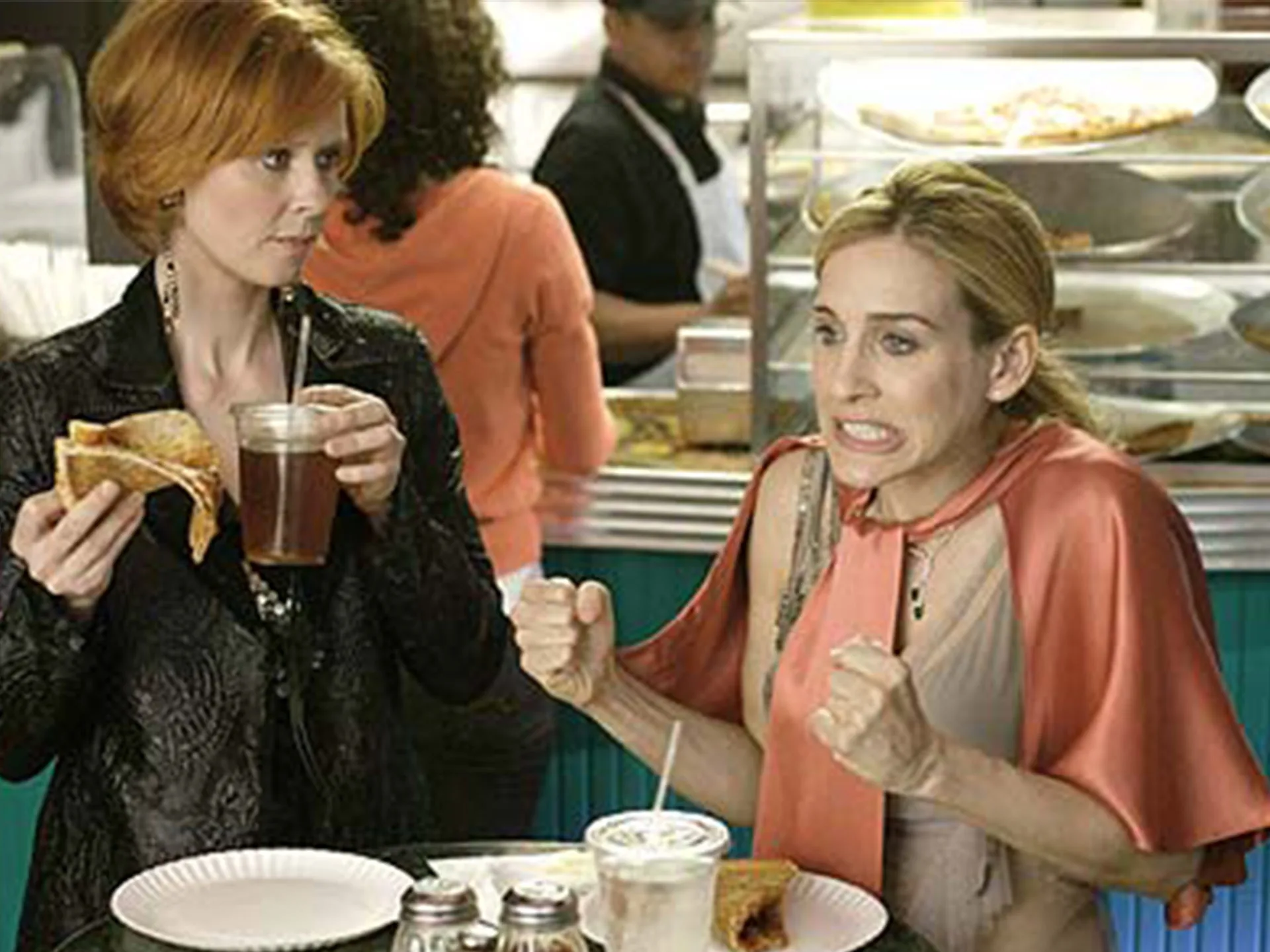Two women in a diner: one holding pizza, the other nervously clenching fists, with pizza display in background.