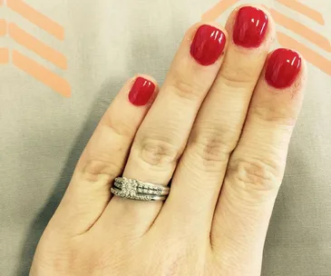 A hand with red nail polish wearing a sparkling engagement ring with a band on the ring finger.