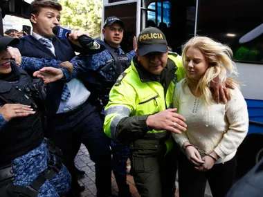 A woman in handcuffs is escorted by police through a crowd of officials and media.