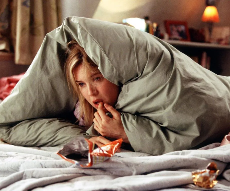 Person bundled in a duvet on a bed, surrounded by snack wrappers, looking pensive.