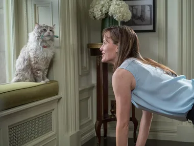 A woman leans forward playfully towards a fluffy cat sitting on a window seat, both inside a well-decorated room.