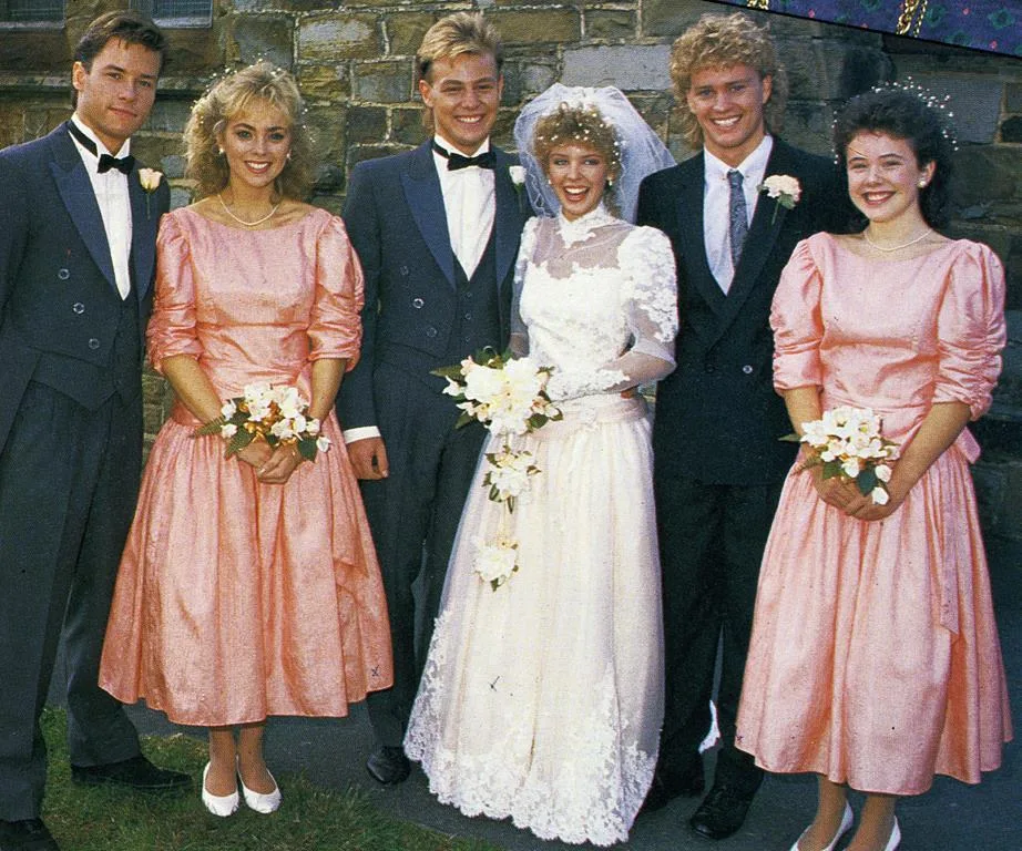 Wedding party in 1980s fashion with bride in white dress and veil, surrounded by bridesmaids and groomsmen in formal attire.