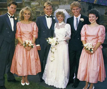 Wedding party in 1980s fashion with bride in white dress and veil, surrounded by bridesmaids and groomsmen in formal attire.