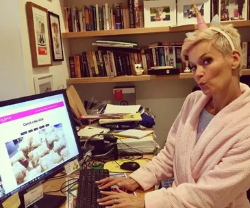 Woman in pink robe with unicorn headband typing at desk, screen shows a carrot cake recipe, books in background.