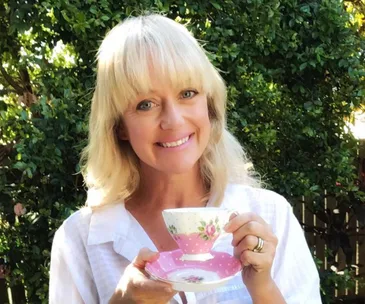 A woman smiling and holding a floral teacup and saucer, with greenery in the background.