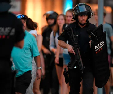 Police officer in tactical gear holding a firearm and shield, with people in the background in a street setting.