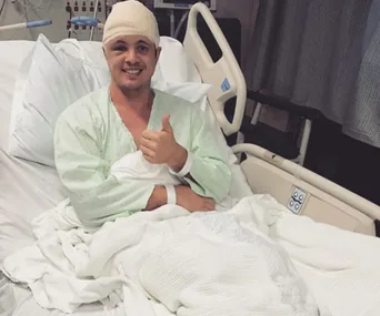"Person in hospital bed with bandaged head, giving thumbs up, wearing a light green hospital gown."