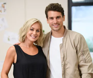 A woman in a black top and a man in a beige jacket smile, standing close together indoors.