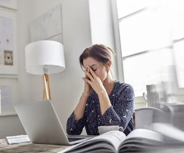 A frustrated woman sitting at a desk with a laptop, holding her head in her hands.