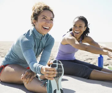"Two women stretching and smiling on the beach, enjoying a sunny day."