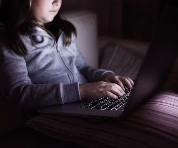A young girl in a grey hoodie using a laptop in a dimly lit room.