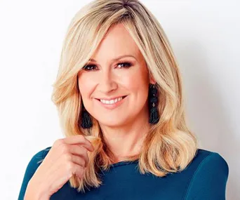 Blonde woman in blue dress smiling, with black tassel earrings, against a plain background.