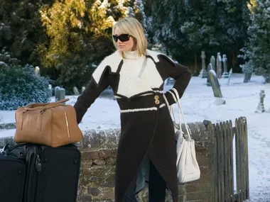 A woman in a stylish winter outfit stands with luggage in a snowy outdoor scene.