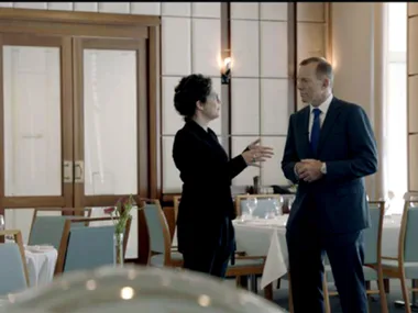 Two people in formal attire are having a conversation in a restaurant with set tables.