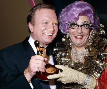 Two people joyfully pose: one with a Logie Award, the other with purple hair and extravagant glasses.