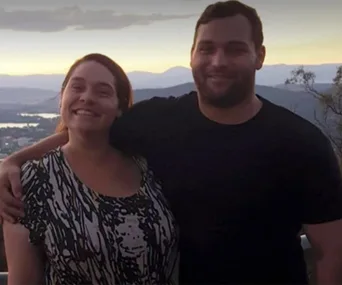 A smiling man and woman standing close with a scenic mountain and sunset background.