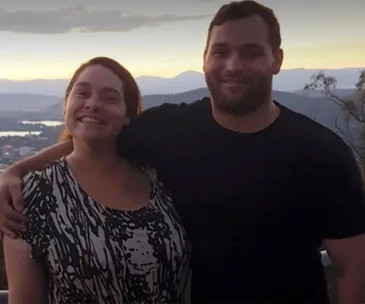 A smiling man and woman standing close with a scenic mountain and sunset background.