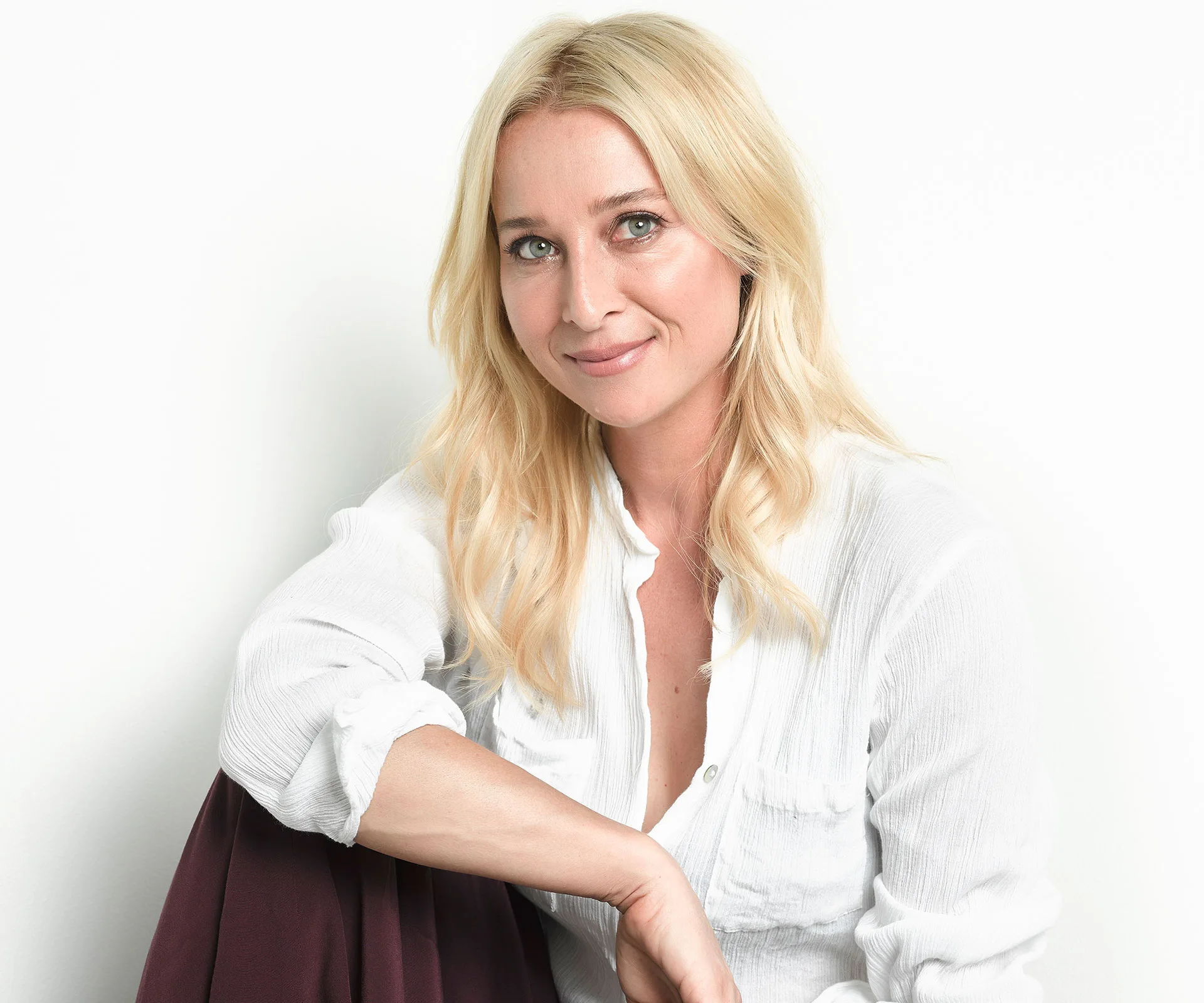 A woman with long blonde hair, wearing a white shirt, smiles softly while seated against a white background.