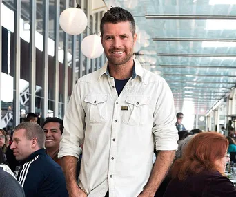 Man in a white shirt smiling at an outdoor restaurant, with a seated crowd in the background under hanging lights.