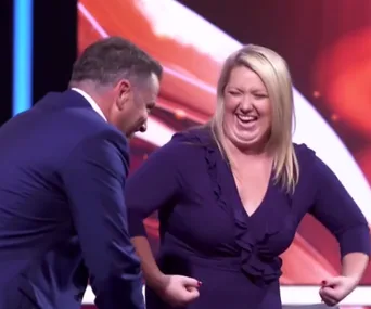 A woman in a blue dress smiling and flexing her arms on a stage with a person in a suit beside her.