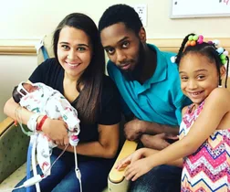 A family of four in a hospital room; a woman holds a newborn baby, with a man and a young girl smiling beside them.
