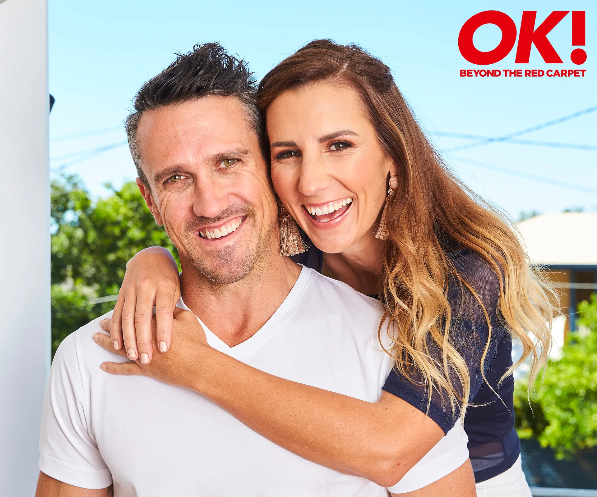 Couple smiling outdoors; woman embraces man from behind; they're casually dressed with greenery in the background.