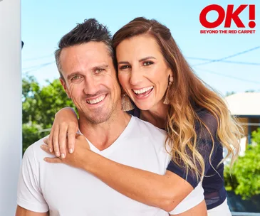 Couple smiling outdoors; woman embraces man from behind; they're casually dressed with greenery in the background.