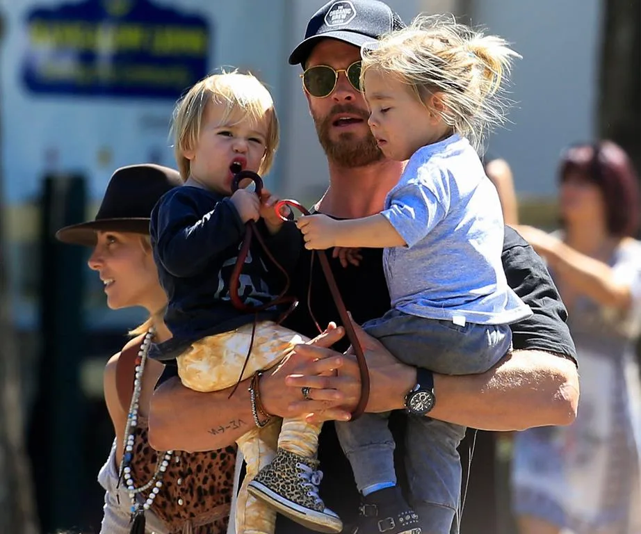 A man carrying two children, one in each arm, outdoors with a woman beside him.