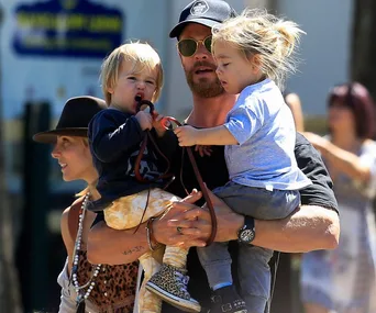 A man carrying two children, one in each arm, outdoors with a woman beside him.