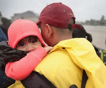A child in a pink hoodie clings to an adult in a yellow raincoat during rescue efforts amidst flooding.