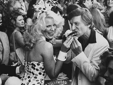 A woman in a polka dot dress and a man in a white suit eat at a crowded outdoor event.