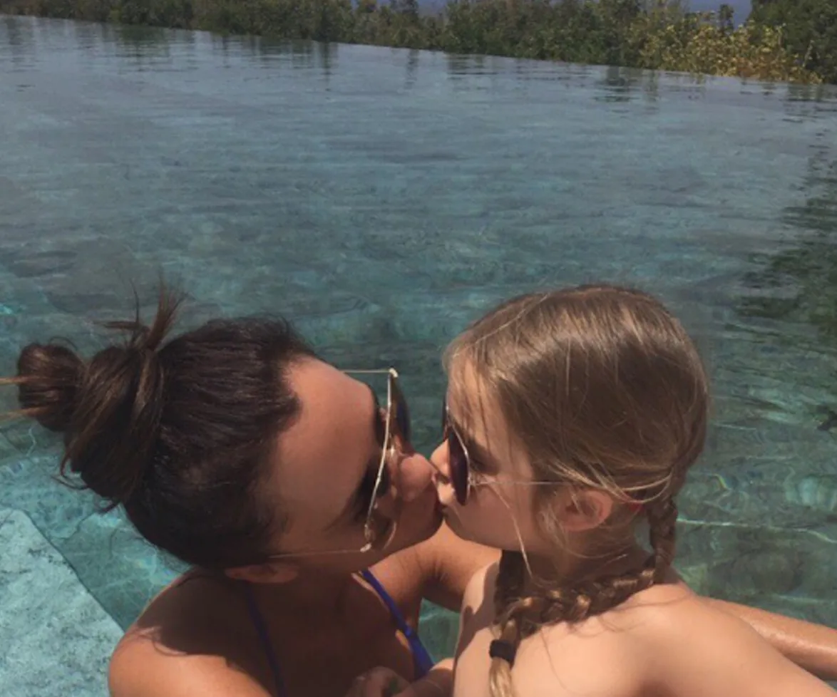 A woman and child wearing sunglasses share a kiss in a swimming pool with clear water and trees in the background.