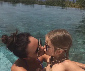 A woman and child wearing sunglasses share a kiss in a swimming pool with clear water and trees in the background.