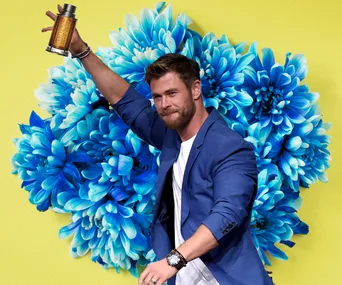 Man in a blue jacket holding a cologne bottle in front of large blue flowers on a yellow background.