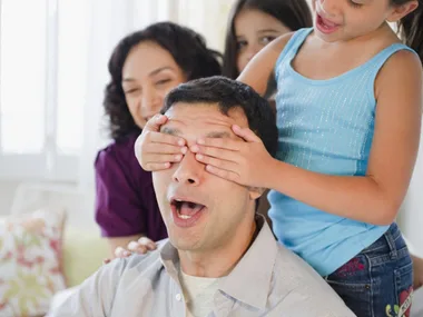 A joyful family moment with a child covering a man's eyes, likely a surprise for Father's Day.
