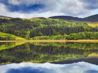 A serene landscape with lush green forests and hills reflecting on a calm lake under a cloudy sky.