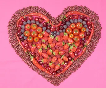 Heart shape made of strawberries, tomatoes, cherries, beans, and peppers on a pink background.