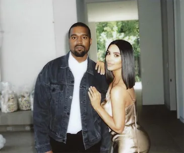 A man in a denim jacket and a woman in a satin dress pose together indoors.