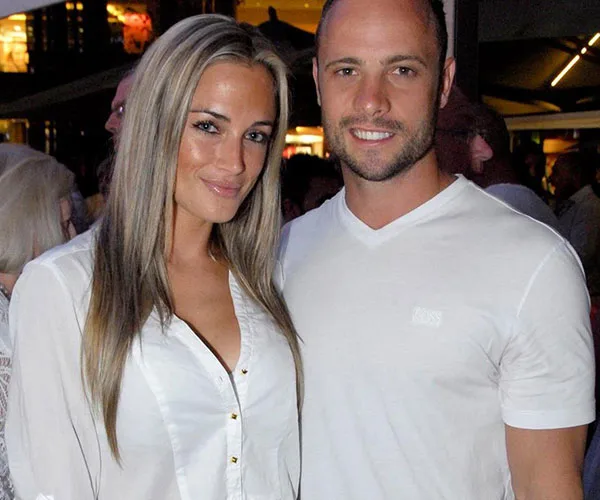 A smiling couple in casual attire, standing closely in a crowded, dimly-lit area.