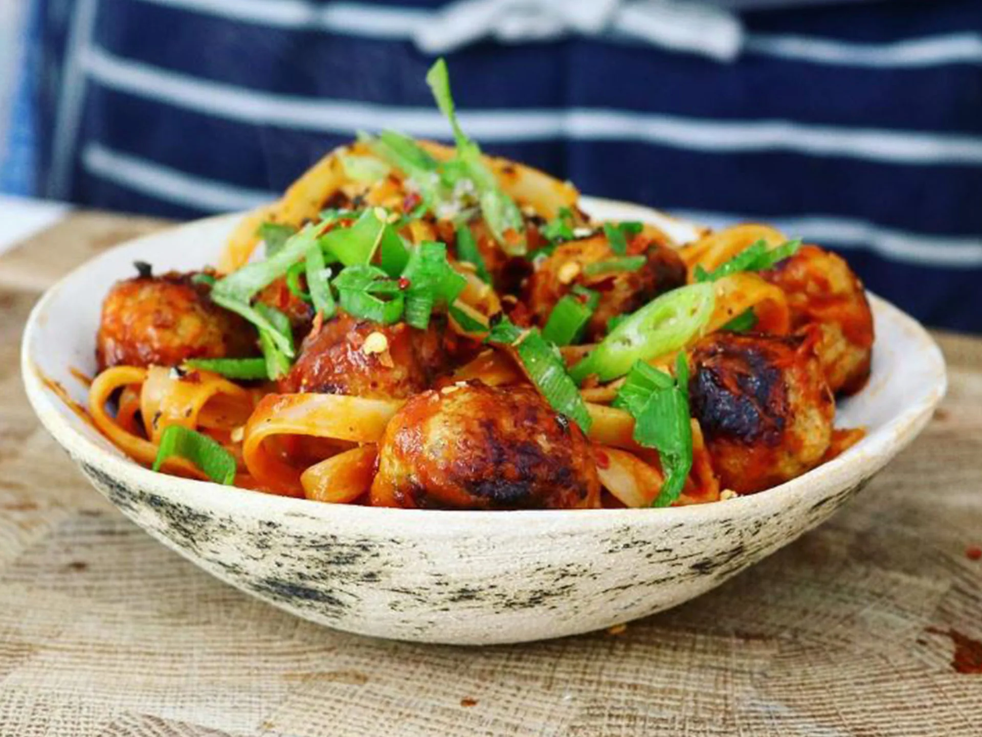 A bowl of pasta with meatballs, topped with green onions and chili flakes on a wooden table.