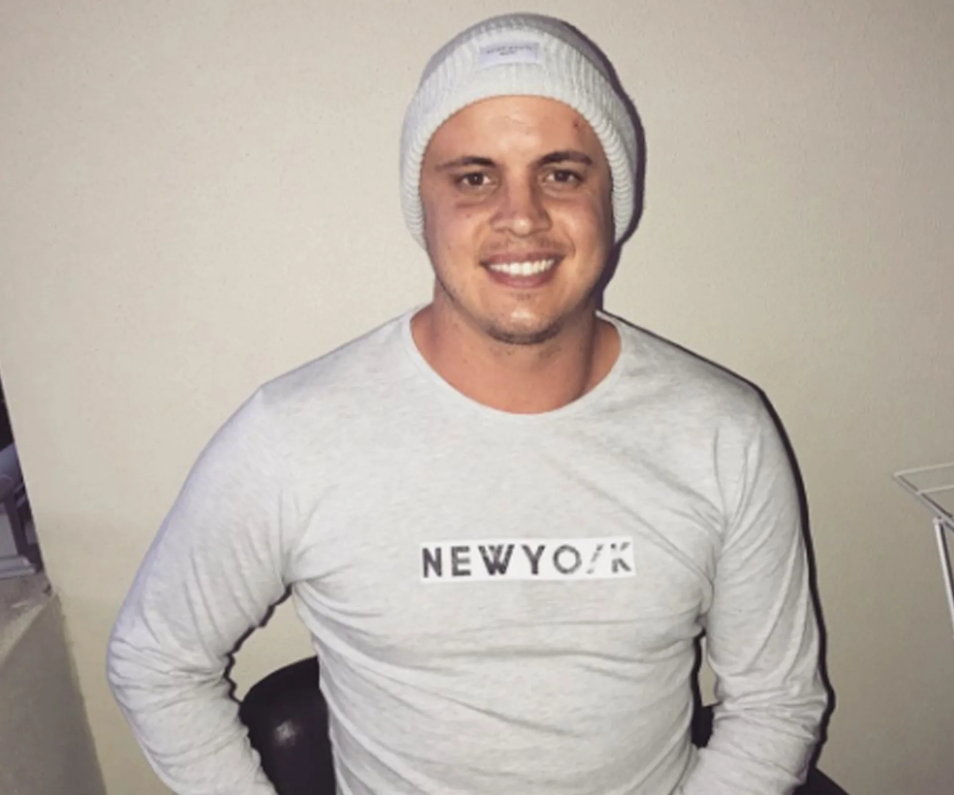 Man in a white beanie and "New York" shirt smiling, seated indoors.
