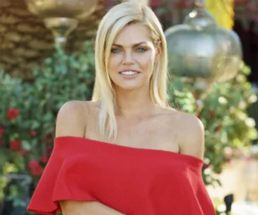 A woman in a red off-shoulder dress stands smiling in a garden setting with a blurred background.