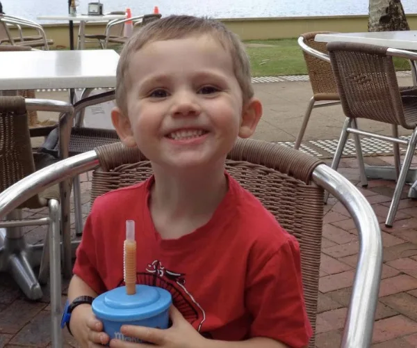 A young child smiling, seated outdoors at a cafe, holding a blue cup with a straw, near a waterfront.