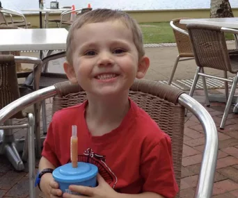 A young child smiling, seated outdoors at a cafe, holding a blue cup with a straw, near a waterfront.