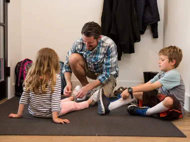 A man helps a young girl with laces while a boy sits nearby; they are in a hall with coats and backpacks.